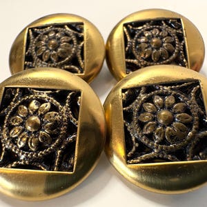 Peut inclure: Quatre boutons circulaires avec un bord en métal doré brossé lisse. Chaque bouton présente un centre carré en retrait contenant un motif floral en relief de couleur bronze oxydé foncé avec une fleur centrale entourée de filigrane.