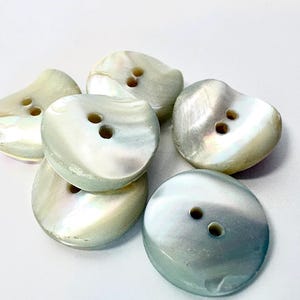 May include: A collection of six irregular, rounded buttons crafted from iridescent mother-of-pearl. Each button features a smooth, polished surface with a natural pearlescent sheen and two central holes for attachment, displaying varied shades of white, grey, and soft blue.