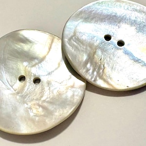 May include: Two round, flat buttons crafted from iridescent mother-of-pearl shell. Each button features a smooth, polished surface with a natural pearlescent sheen and two centred holes for attachment. The material displays subtle white and cream tones.