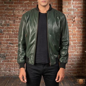 May include: A dark green leather bomber jacket with a black ribbed collar, cuffs, and hem. The jacket has a front zipper closure and two side pockets. The model is wearing a black t-shirt and black jeans. The background is a brick wall.