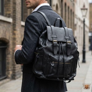 May include: Black leather backpack with a flap closure, drawstring, and two front zippered pockets. The backpack has adjustable shoulder straps and gold-colored buckles. The bag is worn over the shoulder.