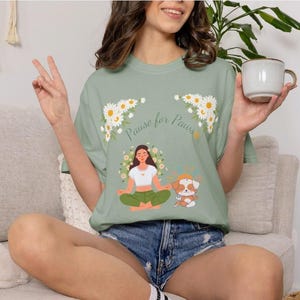 May include: Sage green t-shirt with the text "Pause for Paws" and a graphic of a woman meditating with a dog. The shirt features daisy accents and is made of a soft material. The shirt is a casual wear item.