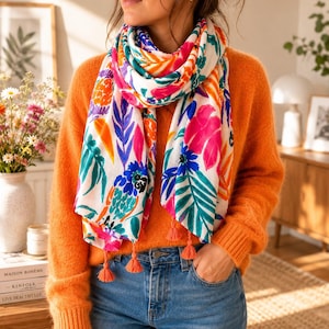Puede incluir: Bufanda blanca con un vibrante estampado floral en rosa, azul, naranja y verde, acentuada con borlas naranjas. La bufanda se lleva con un suéter naranja y vaqueros azules.