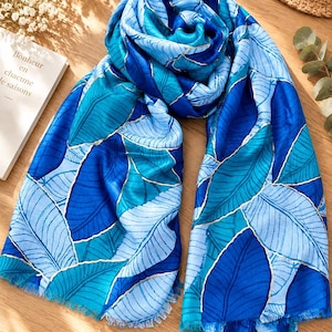 Op de afbeelding: Een rechthoekige sjaal met een levendig patroon van overlappende bladeren in verschillende blauwtinten, waaronder marineblauw, koningsblauw en lichtblauw. Elk blad is omlijnd met een dunne, metallic gouden lijn. De sjaal heeft een gerafelde randafwerking.