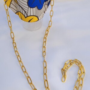 Könnte beinhalten: Ein transparenter Kunststoffbecher mit Deckel und goldenem Strohhalm, ausgestattet mit einer Stoffhülle mit weißem, gelbem und blauem Blumenmuster. Die Hülle hat goldene Metallösen, die an einer langen, strukturierten goldenen Metallkette befestigt sind.