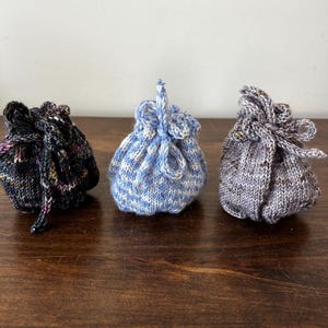 May include: Three small, hand-knitted drawstring pouches. The left pouch is black with multicolored flecks, the center pouch is light blue and white striped, and the right pouch is a heathered grey with brown speckles. Each has a tied top closure.