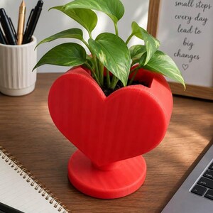 May include: A bright red heart-shaped planter with a circular base, containing a green pothos plant with variegated leaves. The planter has a smooth, matte finish and is positioned upright, showcasing a distinct heart silhouette and a central opening.