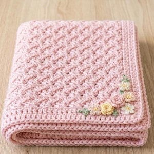 May include: A soft pink crocheted baby blanket featuring a textured basketweave stitch pattern throughout the main body. The corner is decorated with a cluster of small, hand-crocheted flowers in shades of pale yellow, cream, and soft green.