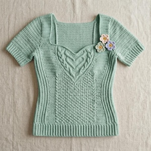 May include: A light sage green short-sleeved crochet top featuring a square neckline, a central heart-shaped cable knit pattern, and three small, multi-colored crocheted flower appliqués attached to the upper right shoulder area.