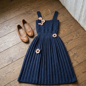 May include: A navy blue sleeveless knit dress featuring a textured bodice, a pleated skirt, and three light pink crocheted flower appliqués. The dress is accompanied by a pair of tan leather ballet flats and a thin gold-tone chain necklace.