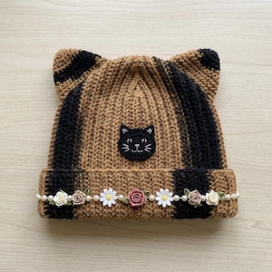 May include: A brown and black striped crochet beanie with cat ears, featuring a black embroidered cat face patch on the front. The folded brim is decorated with a horizontal row of small white pearls, white daisy flowers, and pink roses.