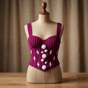 May include: A magenta crochet corset top featuring thick shoulder straps and a sweetheart neckline. The garment is decorated with white embroidered lightning bolt motifs and a vertical row of white pom-poms down the center front panel.