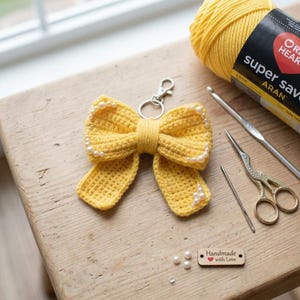 May include: A yellow crochet bow keyring with white pearl embellishments. The bow is attached to a silver clasp. A ball of yellow yarn, crochet hooks, scissors, and a needle are also visible.