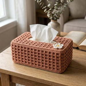 Può includere: Un copriscatola per fazzoletti rettangolare con un motivo all'uncinetto a punto waffle color terracotta. Un piccolo fiore all'uncinetto bianco a cinque petali con il centro giallo è fissato nell'angolo superiore. Un fazzoletto bianco fuoriesce.