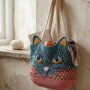 Op de afbeelding: Een gehaakte tas in de vorm van een kattengezicht, met een blauwgroen opengewerkt bovendeel, oren met gele accenten, blauw-gele ogen en een effen oudroze onderkant met een klein roze neusje en donkergrijze geborduurde snorharen.
