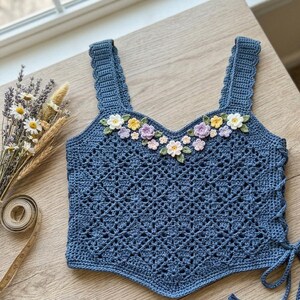 May include: A blue crochet tank top featuring a diamond-patterned openwork design, wide shoulder straps, and a V-neckline adorned with small, multi-colored floral appliques. The sides of the garment include lace-up ties for an adjustable fit.