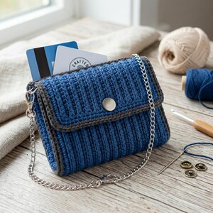 May include: A handmade blue crochet wallet with a silver chain strap. The wallet has a ribbed texture, a gray border, and a silver snap closure. Credit cards are visible inside. Craft supplies, including yarn and a crochet hook, are in the background.