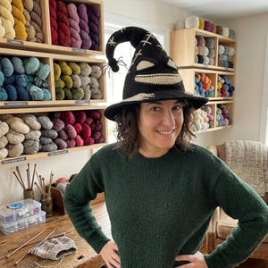 Puede incluir: Un sombrero de bruja de ganchillo negro con una punta curvada y una peque&ntilde;a ara&ntilde;a negra adherida. El sombrero incluye detalles cosidos en color crema que forman una boca ancha y sonriente y ojos, con patrones de punto de cruz en la copa.