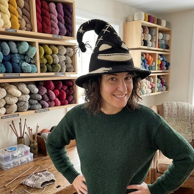 Puede incluir: Un sombrero de bruja de ganchillo negro con una punta curvada y una peque&ntilde;a ara&ntilde;a negra adherida. El sombrero incluye detalles cosidos en color crema que forman una boca ancha y sonriente y ojos, con patrones de punto de cruz en la copa.