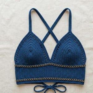 May include: A handmade, dark blue crochet top with a V-neck design and crisscross straps. The top features a gold chain detail along the bottom edge and a tie closure at the back. The crochet work is detailed and intricate.
