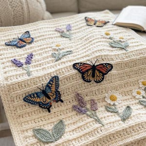 May include: A cream-colored crocheted blanket featuring raised horizontal ridges and appliqued motifs. The design includes various butterflies in orange, black, and blue, alongside sprigs of purple lavender and white daisies with green leaves.