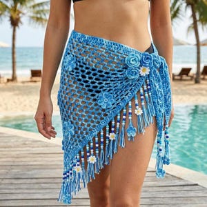 Peut inclure: Un paréo bleu en crochet avec des détails floraux et perlés. L'enveloppe présente un motif en forme de filet, orné de fleurs bleues et de décorations blanches en forme de marguerite. Des glands avec des perles et des fleurs pendent du bord inférieur.