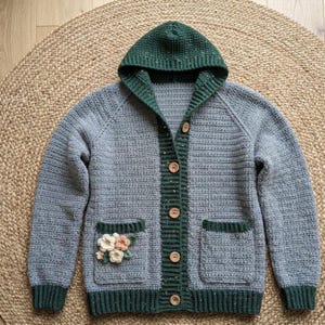 May include: A light gray crocheted cardigan featuring a dark green hood, cuffs, hem, and button placket. The garment has five wooden buttons and two front pockets, with the left pocket decorated with small, crocheted white and peach flowers.