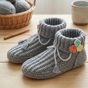 May include: A pair of gray crocheted slippers featuring a ribbed texture, a folded cuff, and a decorative accent of three small crocheted flowers in peach, yellow, and mint green. Each slipper includes a thin gray cord with a small tassel.