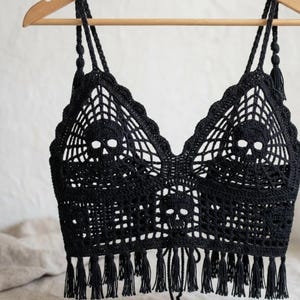 Puede incluir: Un top corto de ganchillo negro con un patrón de calavera detallado integrado en un diseño de telaraña. La prenda tiene tirantes finos, un escote festoneado y un dobladillo inferior acabado con una fila de borlas de flecos negros.