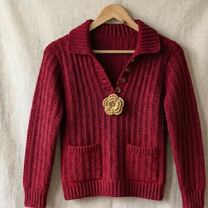 May include: A deep red long-sleeve sweater featuring a ribbed knit texture, a collared neckline with a partial button placket, two front patch pockets, and a decorative gold-toned crocheted flower brooch pinned to the center of the chest.