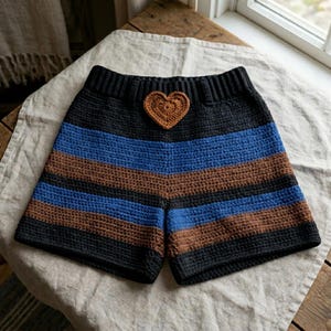 May include: Crochet shorts featuring a high-waisted ribbed waistband and horizontal stripes in black, blue, and brown. A small, textured brown crochet heart is centered at the front of the waistband. The garment is crafted with a consistent stitch.