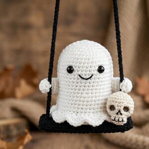 May include: A white crocheted ghost figure with black bead eyes and a stitched smile, sitting on a black crocheted swing. The ghost holds a small, beige crocheted skull in one hand, and the swing is suspended by two black crocheted ropes.