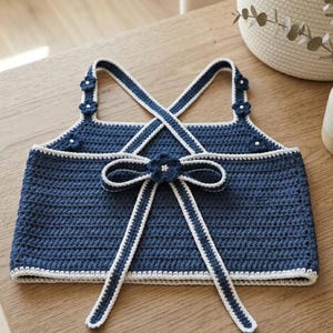 May include: A dark blue crochet crop top featuring white trim along the edges, crisscross straps, and decorative dark blue flower accents. The back of the garment includes a central dark blue bow detail with a small white flower at its center.