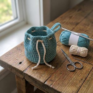 May include: A teal crochet drawstring bag with a handle, sitting on a wooden surface. A ball of teal yarn, a crochet hook, scissors, and a small ball of beige yarn are next to the bag. The bag has a beige drawstring.