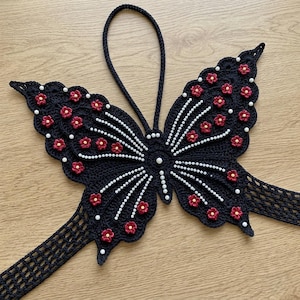 May include: A black crocheted butterfly ornament featuring a top hanging loop and a wide band extending from the lower wings. The butterfly is decorated with small red flower appliques with yellow centers and white beads arranged in radiating lines.