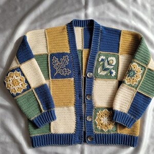 Könnte beinhalten: Eine langärmelige Häkelstrickjacke mit einem Patchwork-Design aus cremefarbenen, senfgelben, salbeigrünen und marineblauen Quadraten. Das Kleidungsstück hat vier dunkle Knöpfe, einen gerippten marineblauen Saum sowie gestickte Blumen- und Blattmotive.