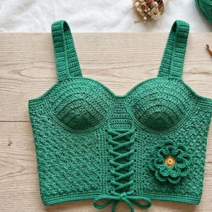 May include: A bright green crochet crop top featuring a corset-style lace-up front with matching green cord ties. The garment has thick shoulder straps, molded cups, and a textured stitch pattern, accented by a single large green flower with a yellow center.