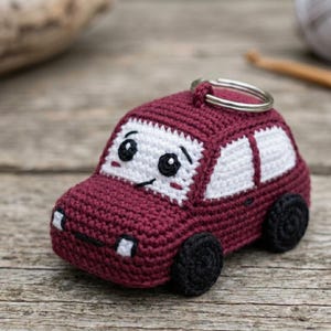 May include: A maroon crochet car keychain featuring a white windshield with black embroidered eyes and a small smile. The vehicle has white side windows, black wheels, and two small white front headlights. A silver metal ring is attached to the top.