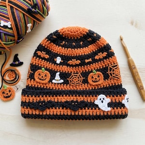 May include: A crocheted beanie featuring horizontal stripes of black and orange yarn. The hat is decorated with small appliqués including orange pumpkins, white ghosts, black bats, white witch hats, and spider webs in a repeating pattern.