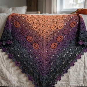May include: A triangular crochet shawl featuring a gradient color transition from orange at the top to purple and dark gray at the bottom. The shawl is decorated with small, raised pumpkin motifs integrated into the intricate, lacy stitch pattern.