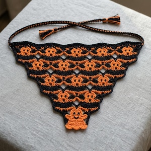 May include: A triangular crochet bandana featuring a repeating pattern of orange ghosts on a black background. The accessory has a scalloped bottom edge and includes two long braided ties with small orange and black tassels at the ends.