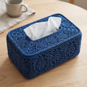 Op de afbeelding: Een rechthoekige tissueboxhoes gemaakt van donkerblauw garen in een gedetailleerd haakpatroon. De hoes heeft een centrale opening waar een witte tissue doorheen wordt getrokken, met een gestructureerd en ingewikkeld ontwerp.