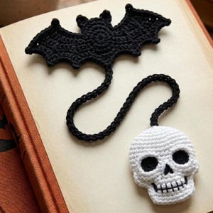 May include: A black crocheted bat bookmark with a long, thin black crocheted cord attached to a white crocheted skull charm. The skull features black yarn detailing for the eye sockets, nose cavity, and teeth, creating a distinct contrast.