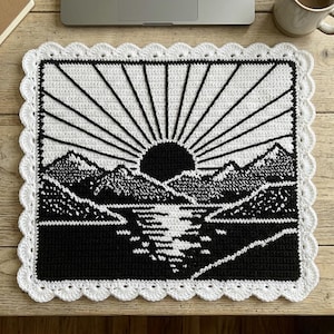 May include: A square, handmade crochet piece featuring a black and white landscape design. The design includes a sunburst, mountains, and a body of water. The border is white with a scalloped edge. This item is suitable for home decor.