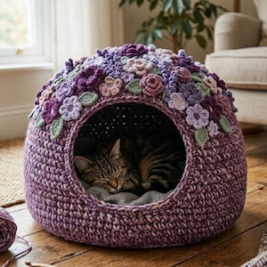 May include: A handcrafted, purple crochet cat bed with a floral design. The bed features a round shape with an opening and is adorned with crocheted flowers in shades of purple, pink, and green. A tabby cat is sleeping inside the bed.