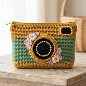 May include: A rectangular crochet pouch designed to resemble a vintage camera. The pouch features mustard yellow and sage green horizontal stripes, a central black circular lens detail with pearl-like beads, and small fabric floral embellishments.