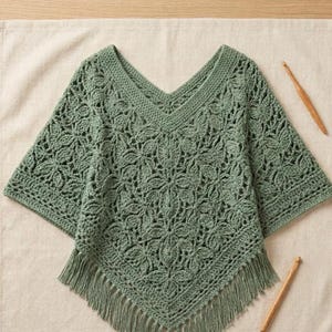 May include: A sage green crochet poncho featuring an intricate floral pattern, a V-neckline, and a fringed hem. The garment is crafted with a detailed openwork stitch design and is accompanied by two wooden crochet hooks placed on the sides.