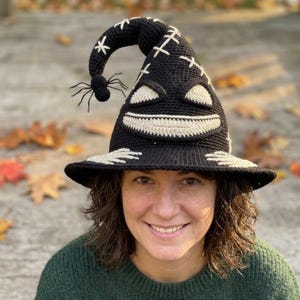 Puede incluir: Un sombrero negro de ganchillo con una parte superior puntiaguda y curva que presenta una ara&ntilde;a negra colgante. El sombrero muestra una cara blanca cosida con dos ojos triangulares y una amplia sonrisa, acentuada por detalles de punto de cruz.