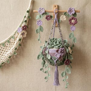 Puede incluir: Colgador de plantas de ganchillo con una cesta colgante llena de una planta suculenta. La cesta está adornada con flores de ganchillo en tonos morado, crema y burdeos. A la izquierda hay una hamaca para plantas de ganchillo a juego.