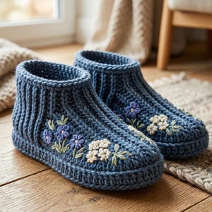 Può includere: Un paio di pantofole blu lavorate a maglia con ricami floreali. Le pantofole hanno un polsino a coste e sono decorate con fiori ricamati viola e bianchi con foglie verdi. Le pantofole sono esposte su una superficie di legno.
